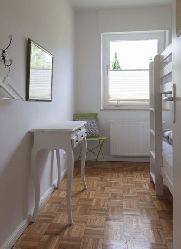A small, well-lit room with a wooden parquet floor, a white radiator, a green chair, a white desk, and a bed with white bedding. - "yachtschule Svaneke" (Gluecksburg)