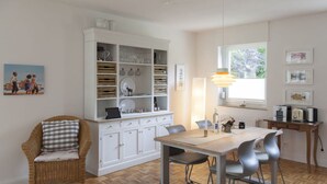 Uma área de refeições com uma mesa de madeira e cadeiras, um armário branco com utensílios de vidro e uma cadeira de vime com almofada xadrez.