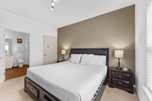 Un dormitorio con una cama, mesitas de noche, una mesilla auxiliar, una puerta y un baño visible al fondo.