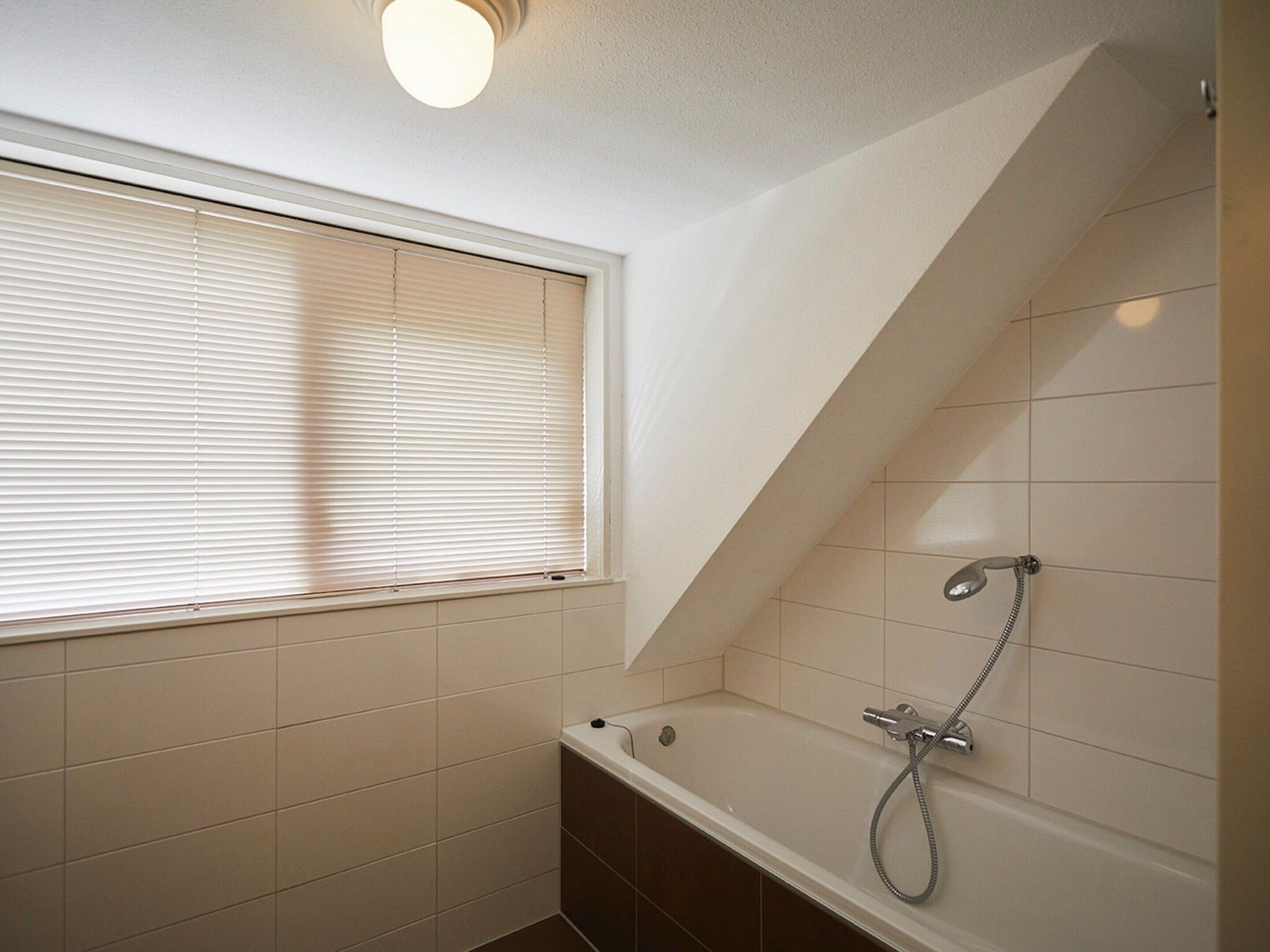 A small, modern bathroom with a bathtub, a shower, and a window with blinds.