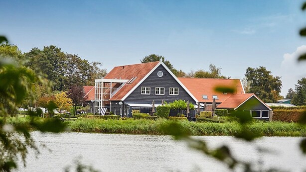 A large, dark-colored building with a red roof, situated by a body of water, surrounded by greenery and trees. - Zeeland Holiday Home Near Beaches (Heinkenszand)