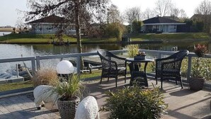 A patio with outdoor furniture, potted plants, and a view of a lake and houses in the background. - Villa Sonnenschein (Makkum)