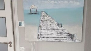 A wall-mounted painting of a wooden pier extending into a calm sea, with a thatched-roof hut at the end. Below the painting, there is a wooden console with a glass candle holder, a small wooden barrel, and a glass pitcher. - Villa Sonnenschein (Makkum)
