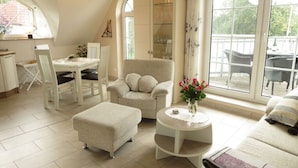 A cozy living room with a sofa, armchair, ottoman, and a small table with a vase of flowers. There is a dining area with a table and chairs, and a large window providing a view of the outdoors. - Apartment Evening sun (Zingst)