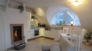 A cozy living room with a fireplace, a dining area with a view through a large window, and a kitchen with modern appliances. - Apartment Evening sun (Zingst)