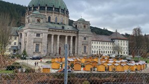 A large historic building with a prominent dome and clock tower, surrounded by a fenced area with small yellow structures. - Meierhof Menzenschwand (St. Blasien)