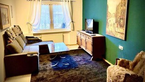 A living room with a sofa, a coffee table, a TV on a wooden cabinet, a painting on the wall, and a window with curtains. - Meierhof Menzenschwand (St. Blasien)