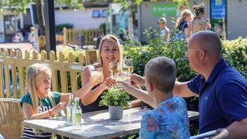 A group of people toasting at an outdoor table with drinks, surrounded by greenery and a wooden fence.