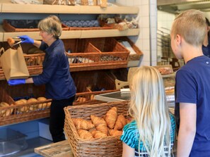 A bakery with shelves stocked with bread and pastries, a person in blue handling a paper bag, and customers waiting.