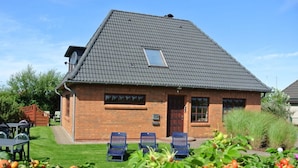 A two-story brick house with a sloped roof, a small window on the second floor, and a front door. There are three blue chairs outside. - South Case in the House aft Diek (St. Peter-Ording)