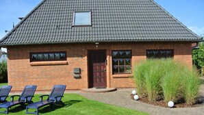 A brick building with a tiled roof, a front door, and a small window. There are three blue chairs in front of the building. - South Case in the House aft Diek (St. Peter-Ording)
