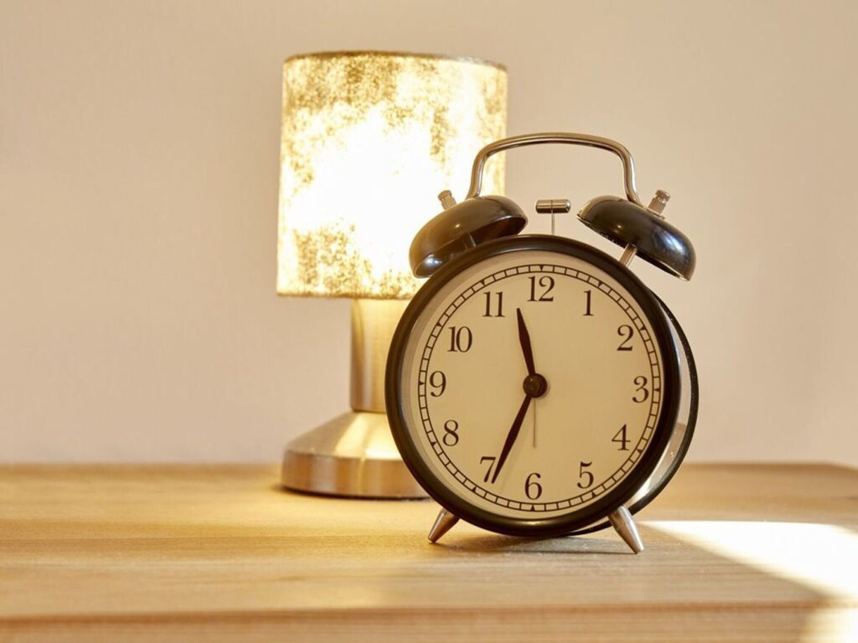 Ein klassischer Wecker mit metallischer Oberfläche und weißem Zifferblatt, platziert auf einer Holzfläche neben einer Lampe mit strukturierter Schirm.