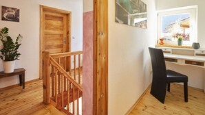 Una escalera de madera conduce a una habitación con un escritorio, una silla, un ventanal y una planta en maceta.
