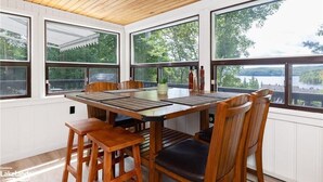 A dining area with a wooden table and chairs, large windows with a view of trees and a lake, and a wooden ceiling.