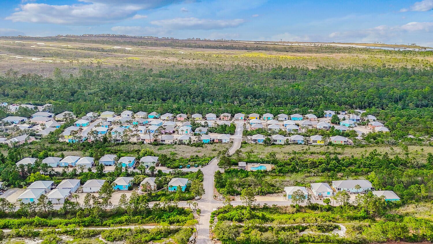 Une vue aérienne d’un quartier résidentiel où les maisons sont disposées selon un plan en forme de grille, entourées de verdure et surplombées par un ciel dégagé.