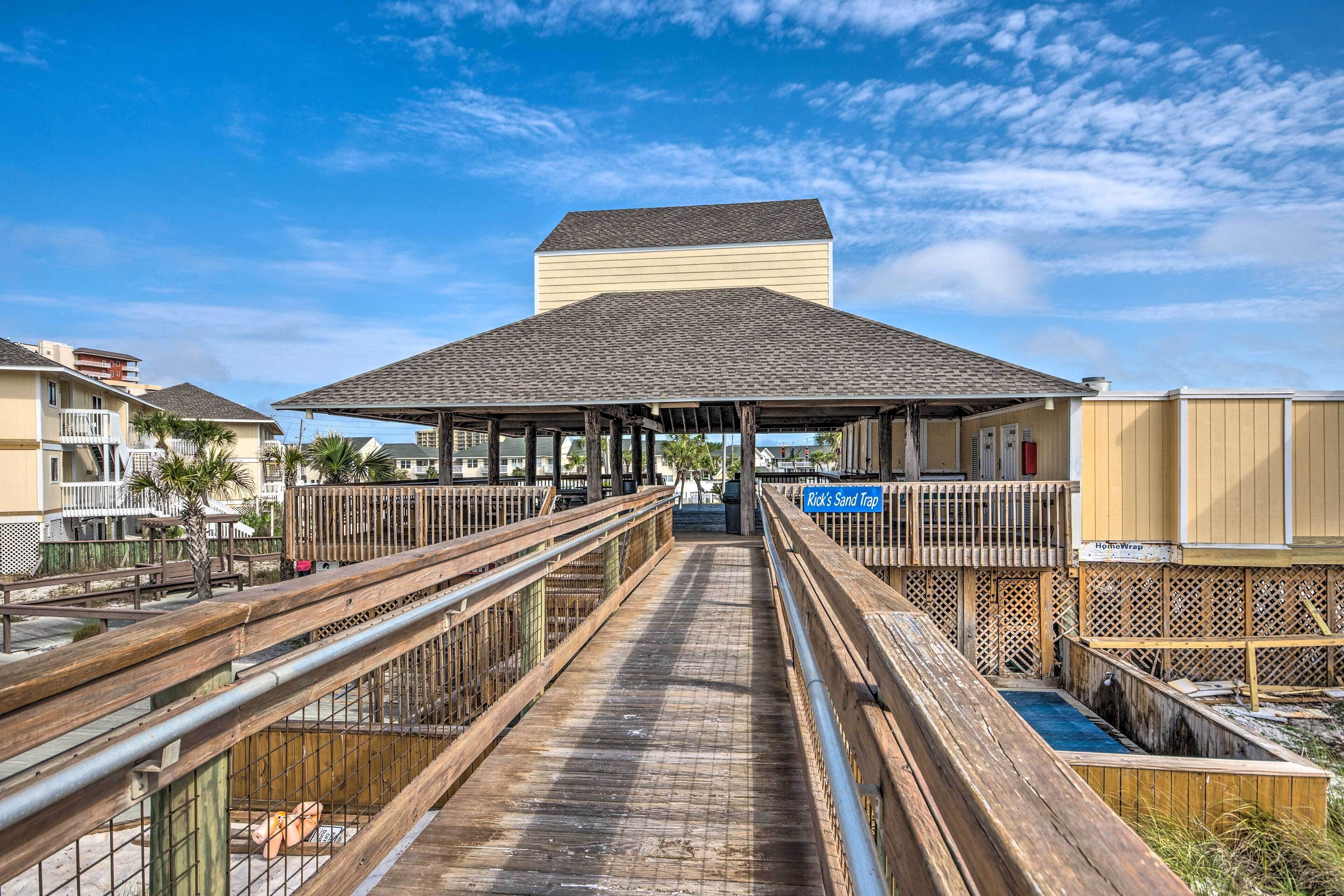 A wooden boardwalk leading to a covered pavilion with a view of a beach and buildings in the background.