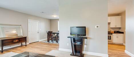 A living room with a glass coffee table, a television on a stand, a dining area with chairs, and a kitchen in the background.