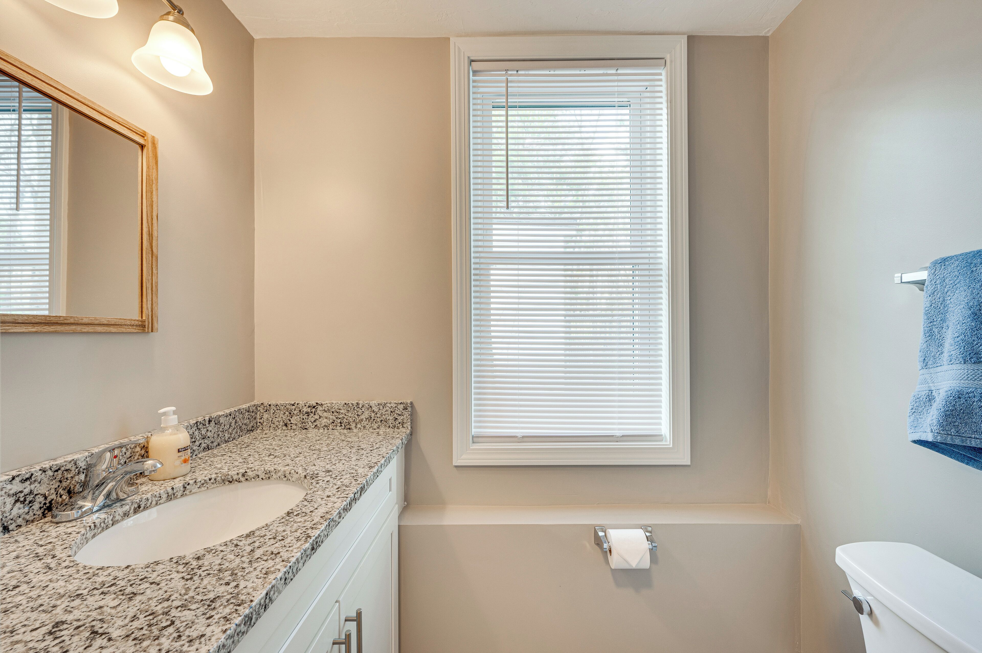 A bathroom with a granite countertop, a sink, a toilet, a window with blinds, and a mirror.