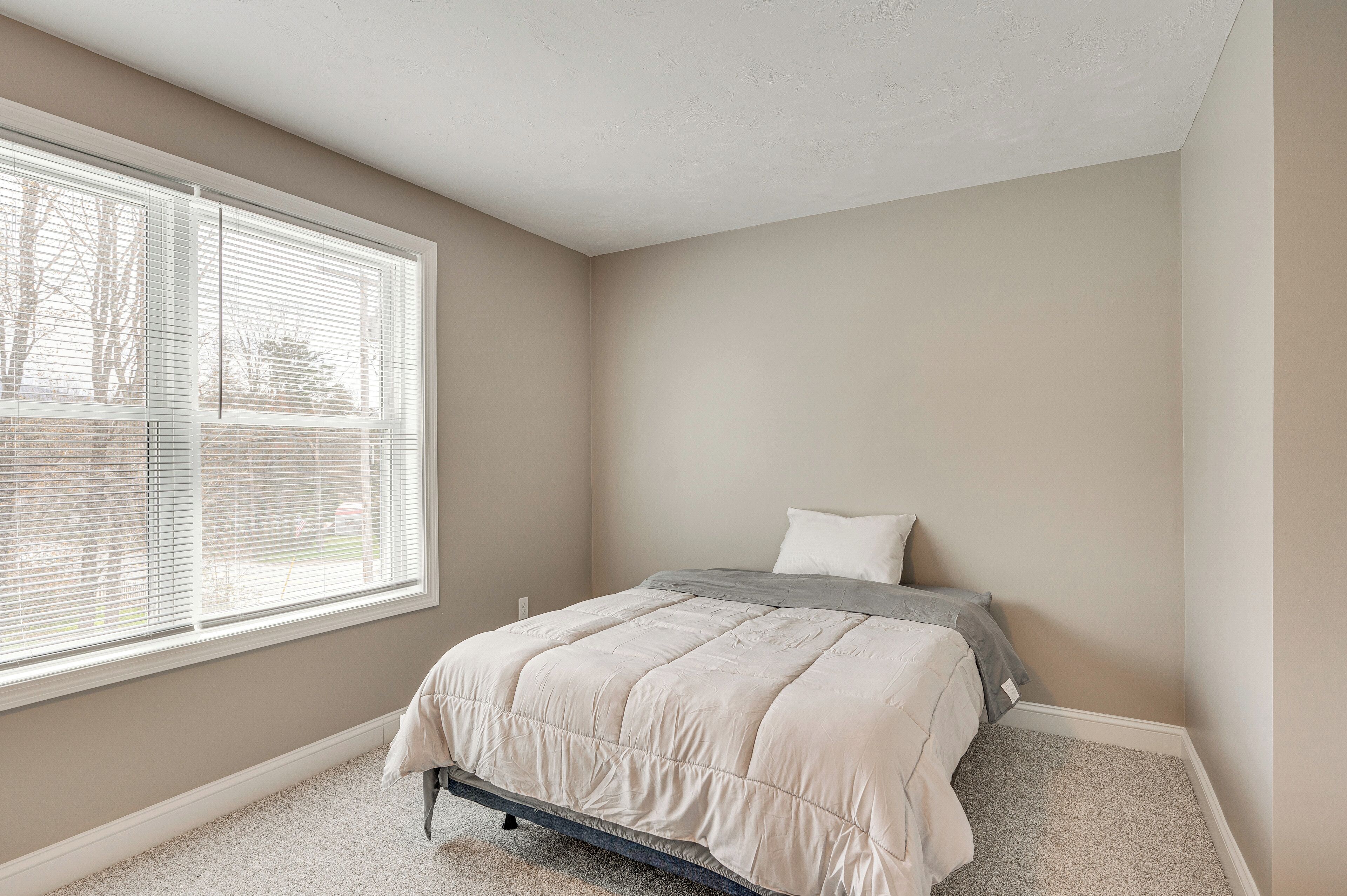 A bedroom with a bed, a window with blinds, and a carpeted floor.