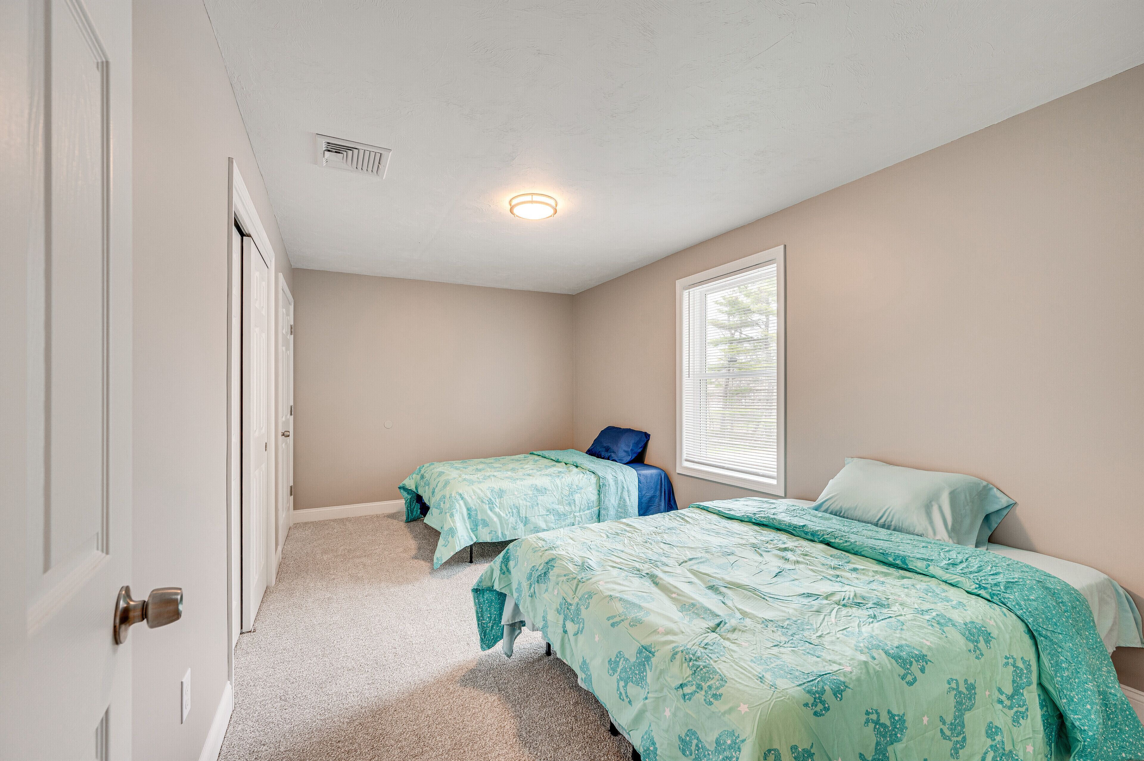 A bedroom with two beds, beige walls, a window, and a door.