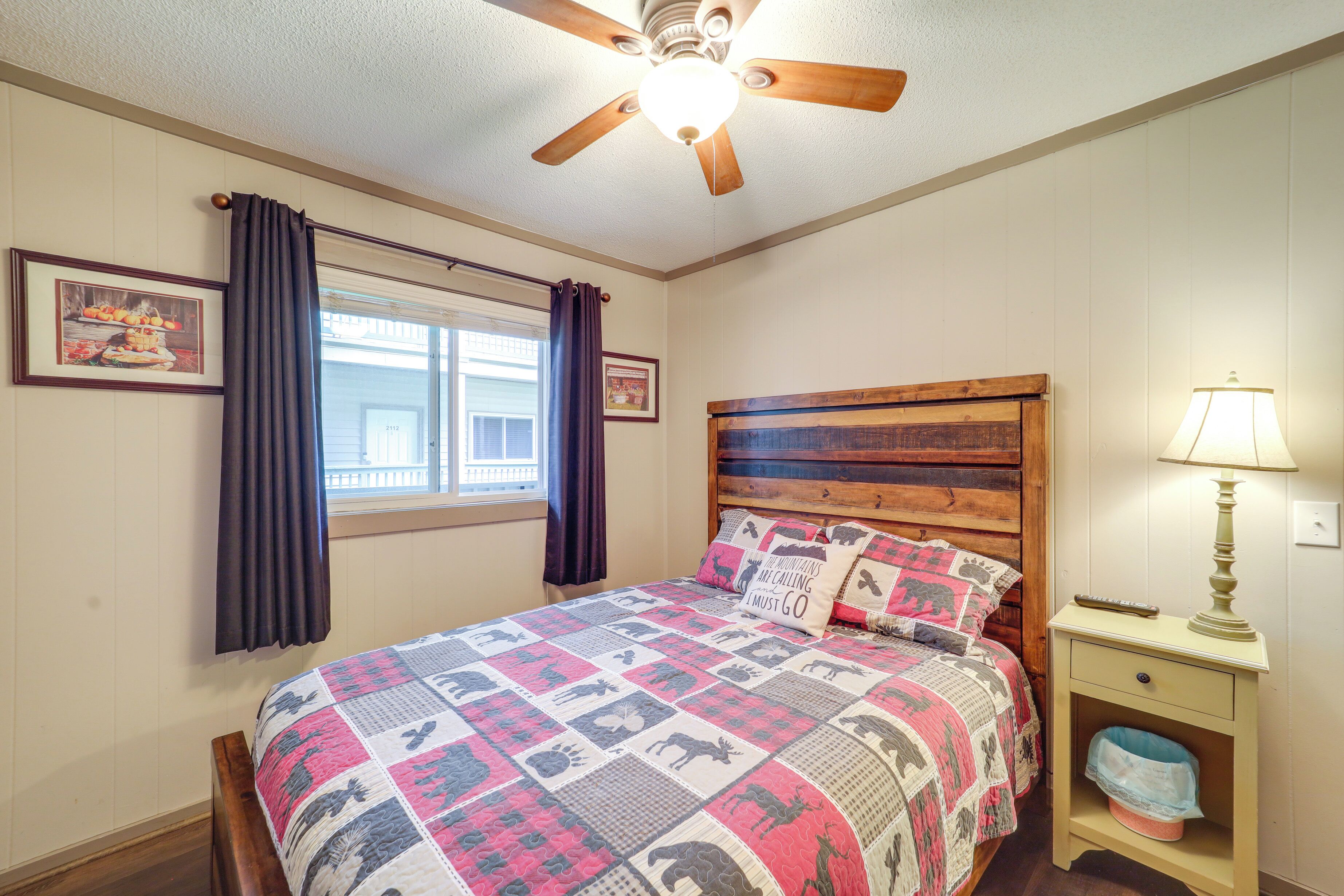 A bedroom with a wooden headboard, a quilted bedspread, a nightstand with a lamp, and a window with curtains.