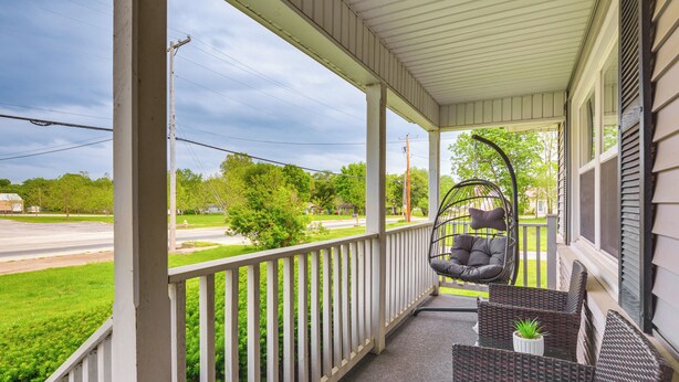 A covered porch with a swing chair and wicker furniture, overlooking a green lawn and street. - Walk to Restaurants: Stockton Home w/ Game Room (Stockton)