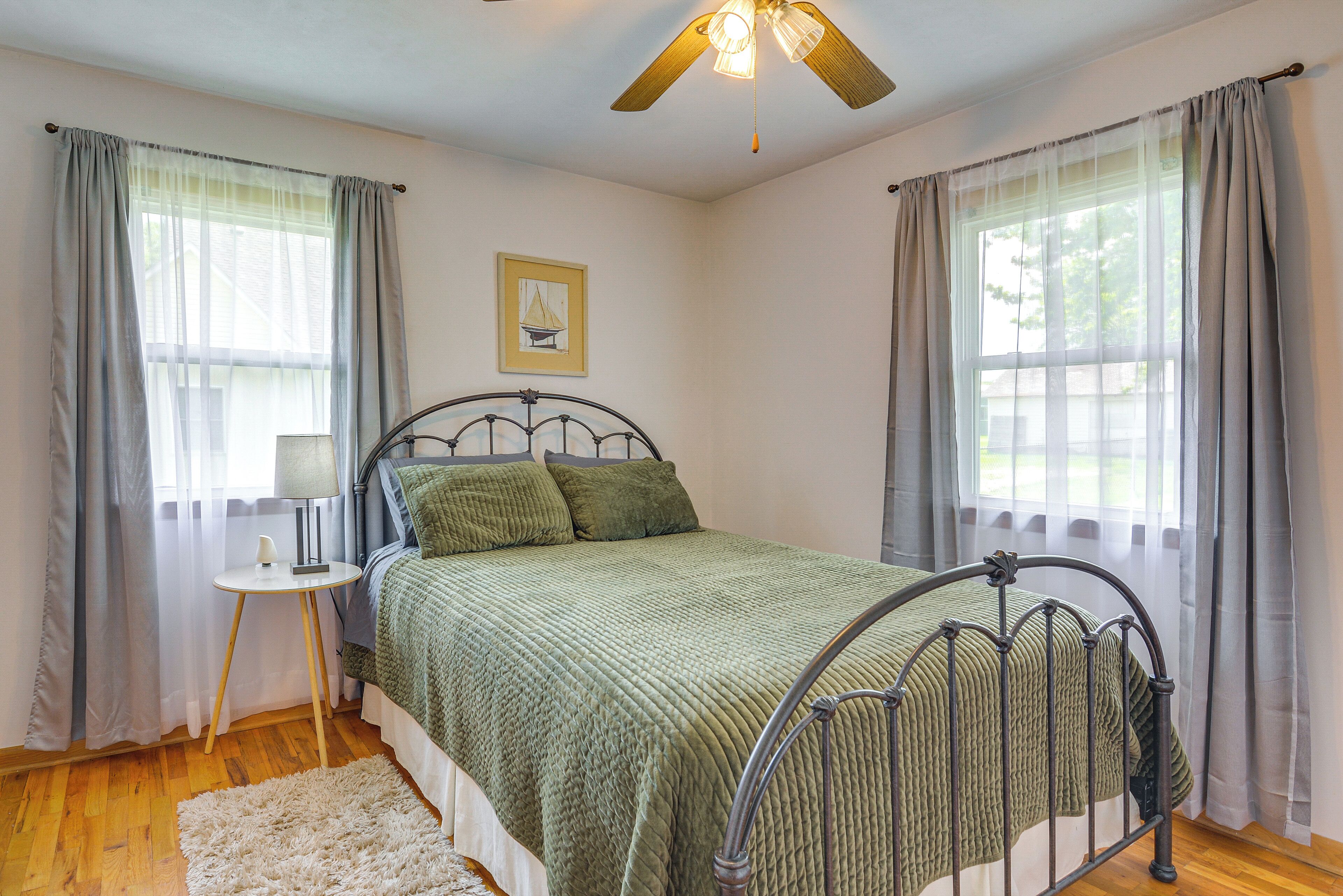 A bedroom with a metal bed frame, a green quilted bedspread, a bedside table with a lamp, and a small rug.