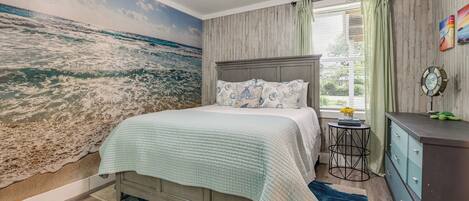 A bedroom with a large ocean mural, a bed with a quilt, a small round table, and a dresser.