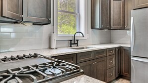 A kitchen with dark wooden cabinets, a stainless steel refrigerator, and a window providing natural light. - Fire Pit + Beach Access: Quiet Northville Retreat (Northville)
