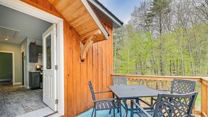 A wooden deck with a table and chairs, a door leading to a kitchen, and a view of trees. - Fire Pit + Beach Access: Quiet Northville Retreat (Northville)
