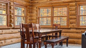 Interieur van een blokhut met een houten eettafel en stoelen, grote ramen met uitzicht op herfstkleuren.