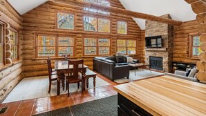Interieur van een blokhut met een eethoek, bank, open haard en grote ramen.