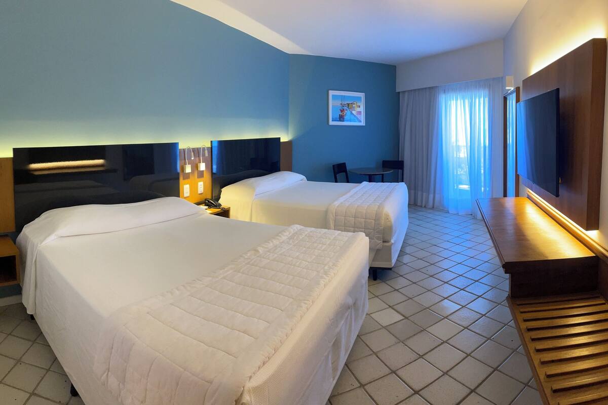 Ein Hotelzimmer mit zwei Betten, einem Schreibtisch, einem Stuhl, einem Fernseher und einem Vorhangfenster.