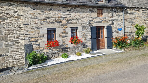 A stone building with two windows and a door, surrounded by gravel and small plants.
