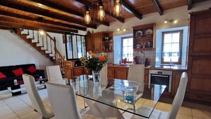 A modern kitchen with a glass dining table, white chairs, wooden cabinets, and a staircase leading to another level.