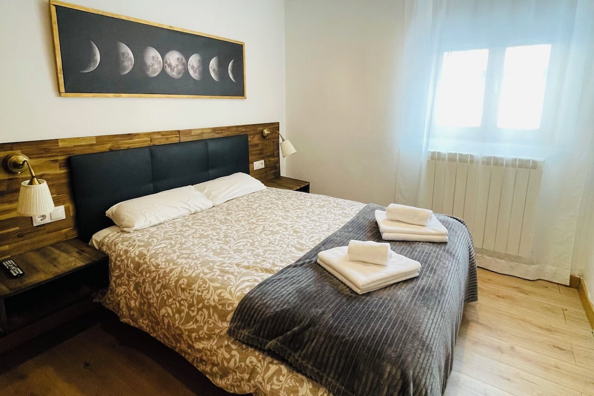Une chambre à coucher bien rangée, avec un lit, des tables de chevet, un tableau accroché au mur et une fenêtre avec des rideaux.