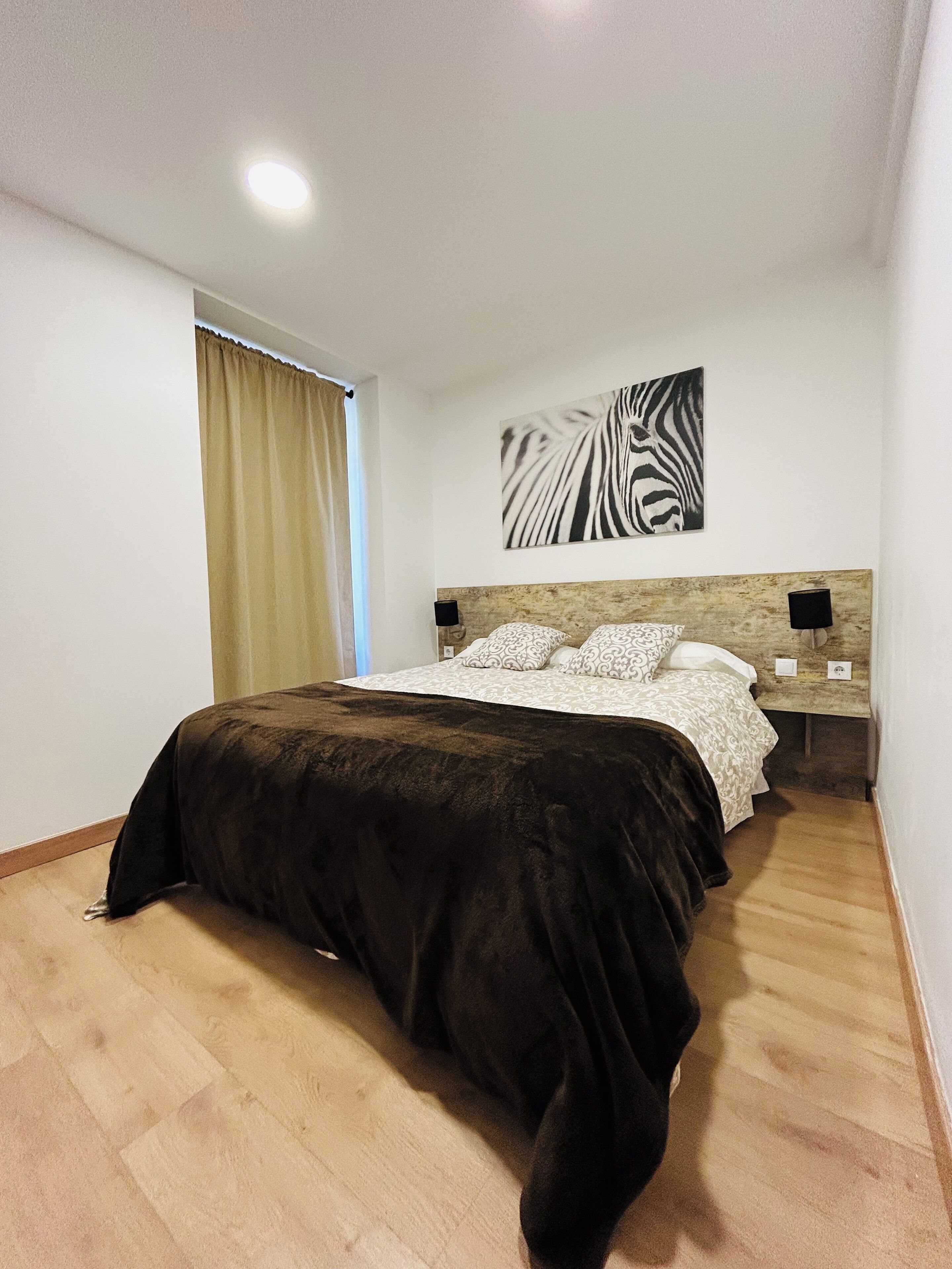 Un dormitorio con cama, cabecero, cuadro de cebra colgado en la pared, mesita de noche con lámpara y ventana con cortinas.