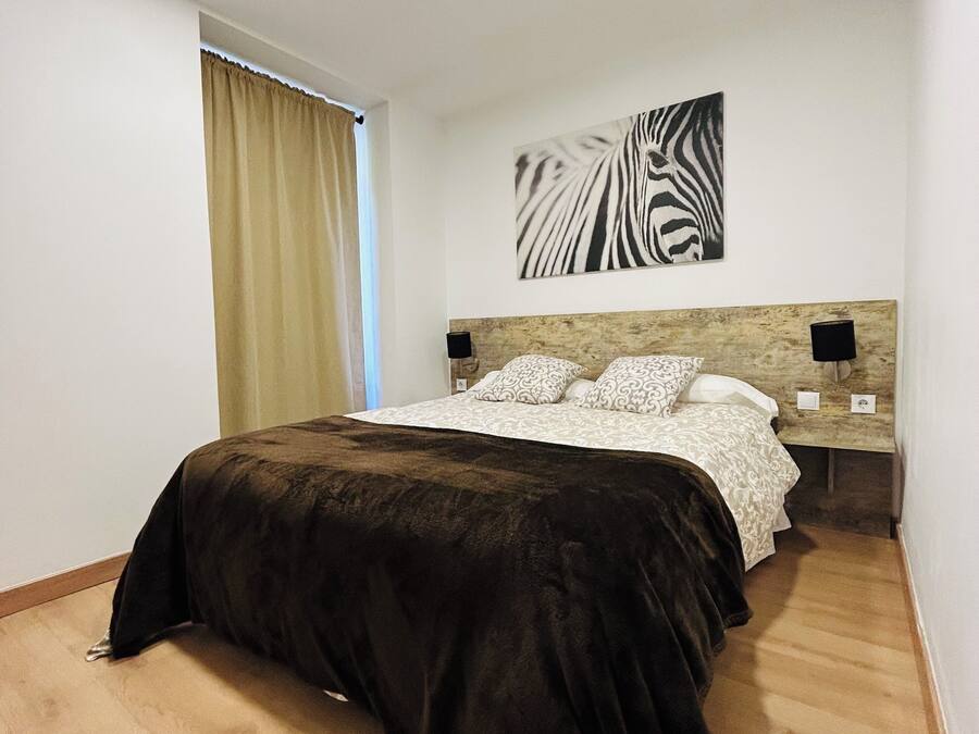 Quarto com cama, cabeceira, quadro com estampa de zebra fixado na parede, mesa de cabeceira com abajur e janela com cortinas.