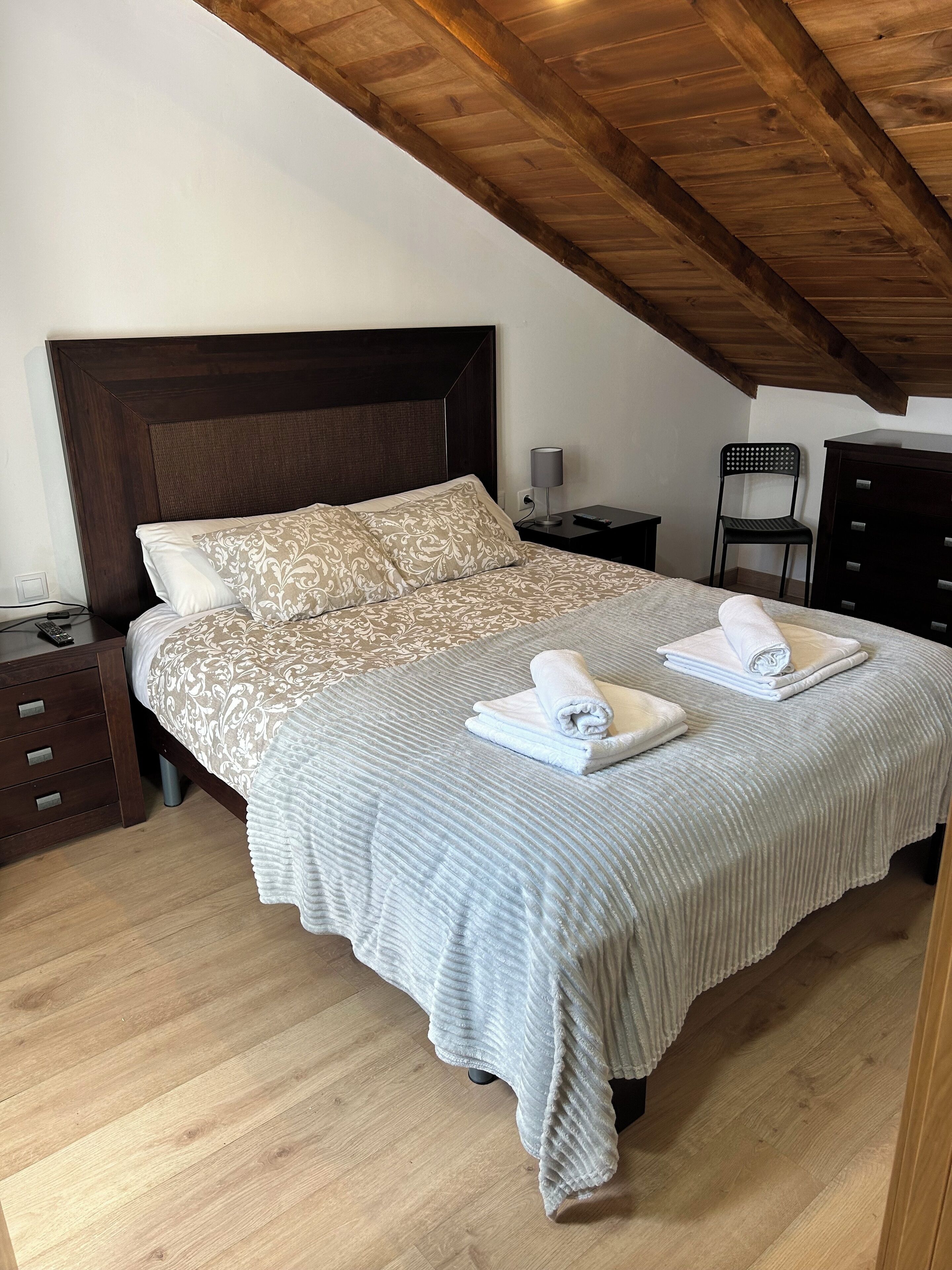 Un dormitorio con techo de madera, una cama con ropa de cama estampada, dos mesitas de noche, una silla y un aparador.