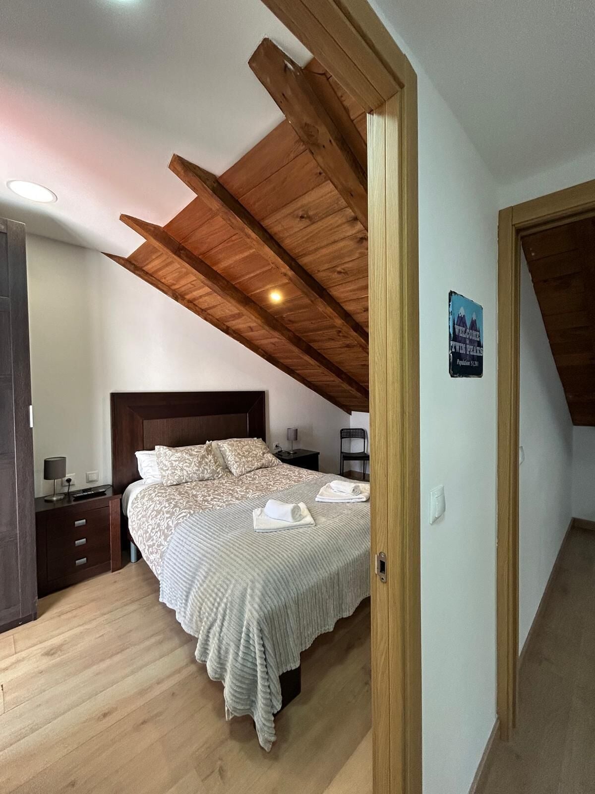 A bedroom with a wooden ceiling, a bed with patterned bedding, a nightstand, and a chair.