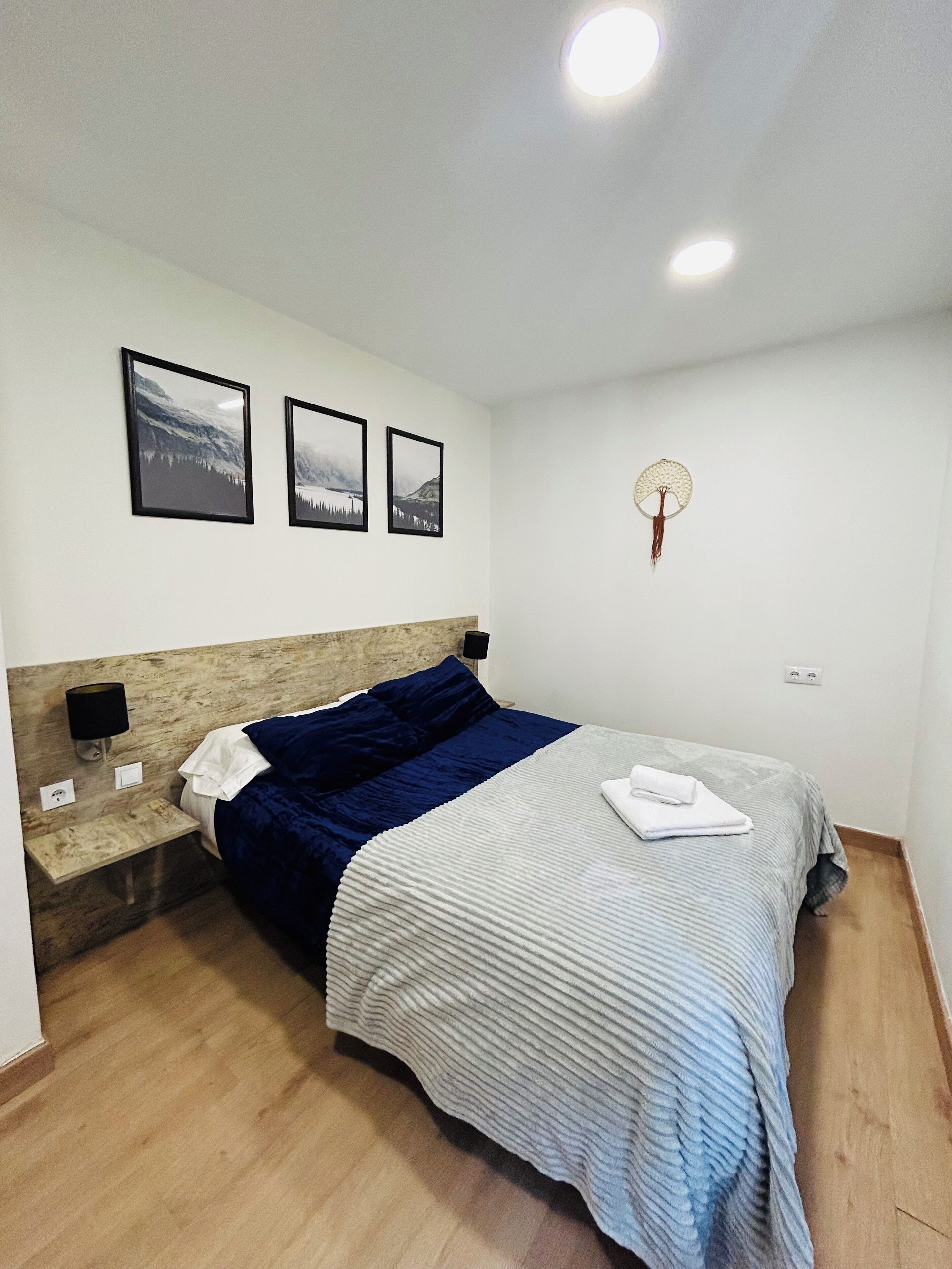 A neatly arranged bedroom with a bed, a nightstand, a wall-mounted lamp, and framed pictures on the wall.