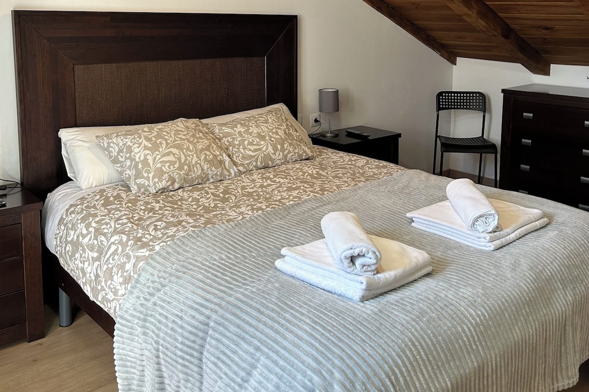Une chambre avec un plafond en pente, un grand lit avec une literie à motifs, une table de chevet, une chaise et une commode.