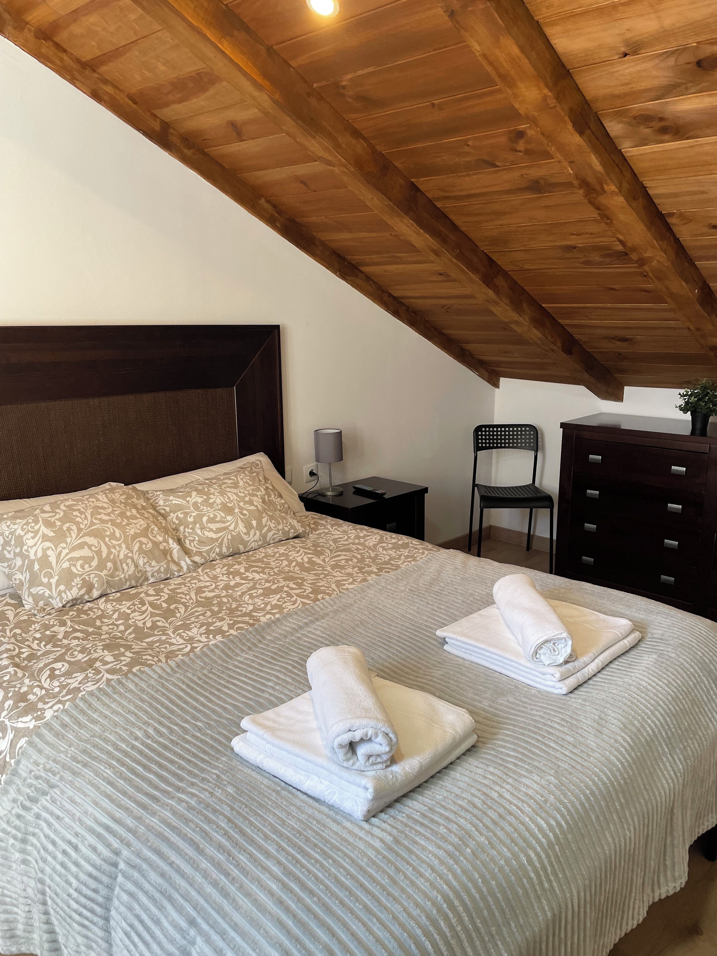 Un dormitorio con techo de madera, una cama con ropa de cama estampada, dos toallas enrolladas, una mesita de noche, una silla y un aparador.