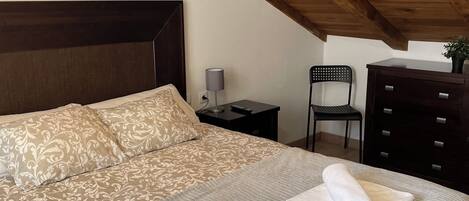 Un dormitorio con techo de madera, una cama con ropa de cama estampada, dos toallas enrolladas, una mesita de noche, una silla y un aparador.