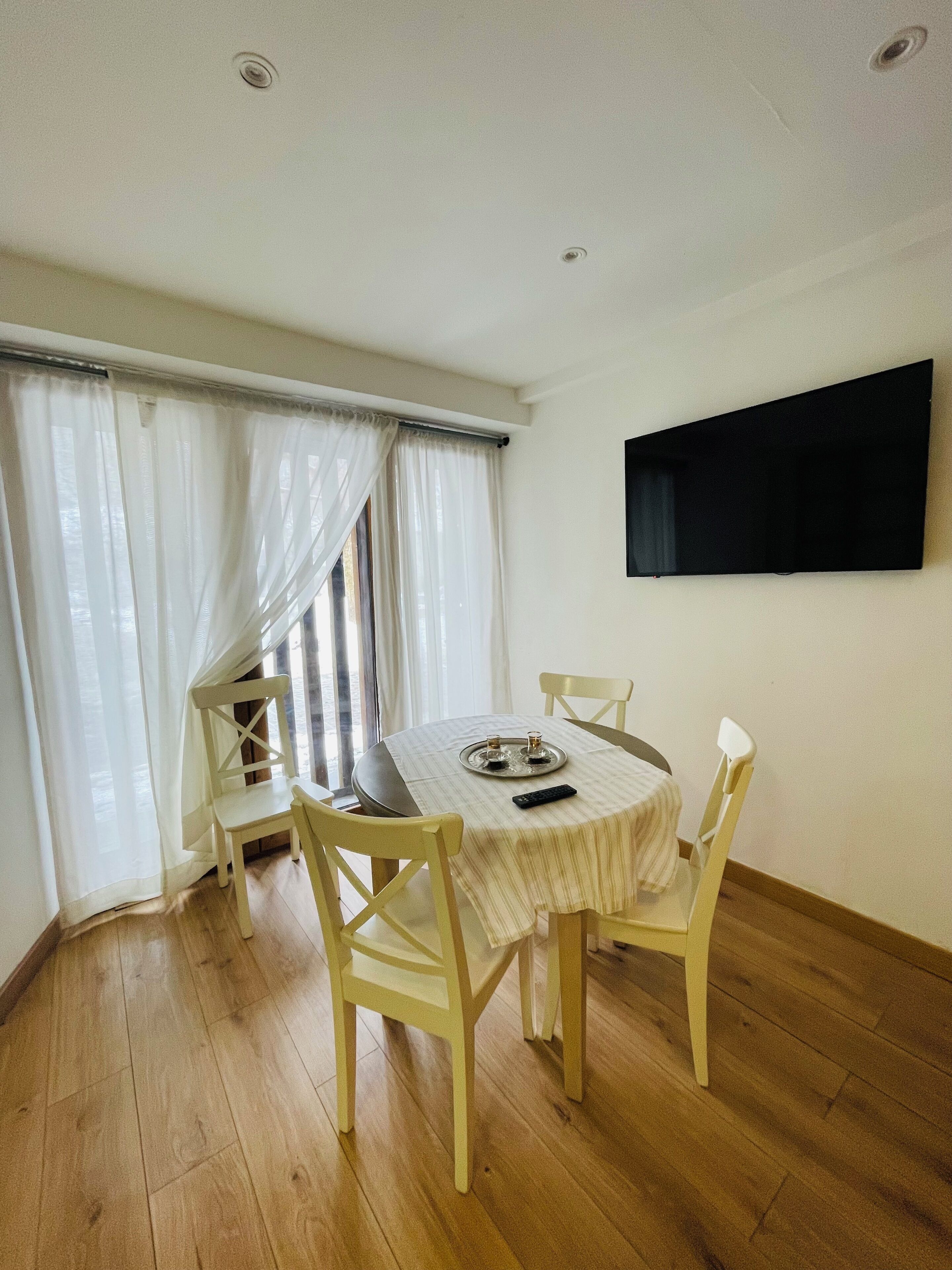 A dining area with a round table, four chairs, a TV mounted on the wall, and a window with sheer curtains.
