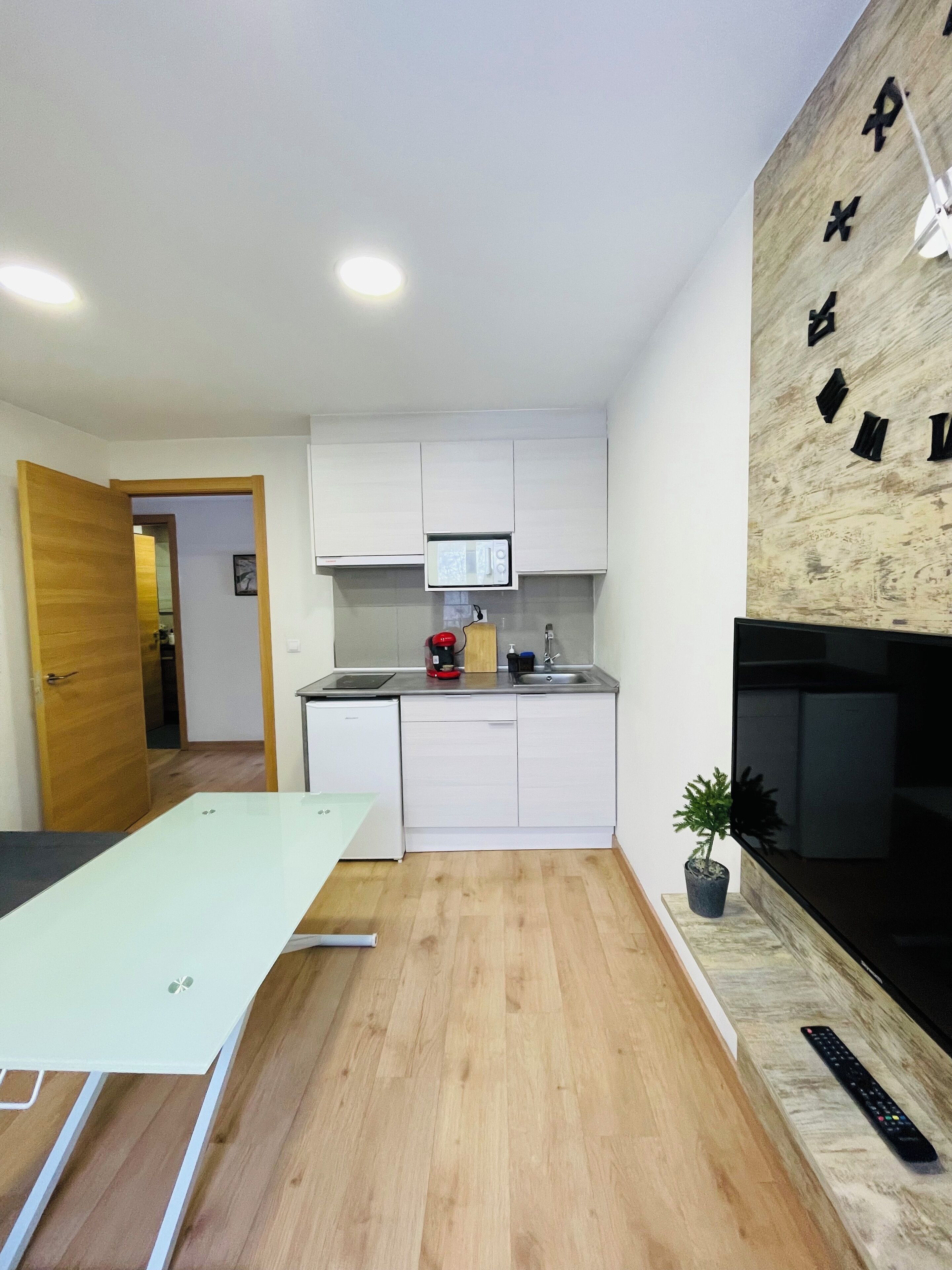 Una cocina moderna con armarios blancos, un microondas, una cafetera y un lavabo. Hay una mesa de comedor con sillas, una planta en maceta y un televisor montado en la pared.