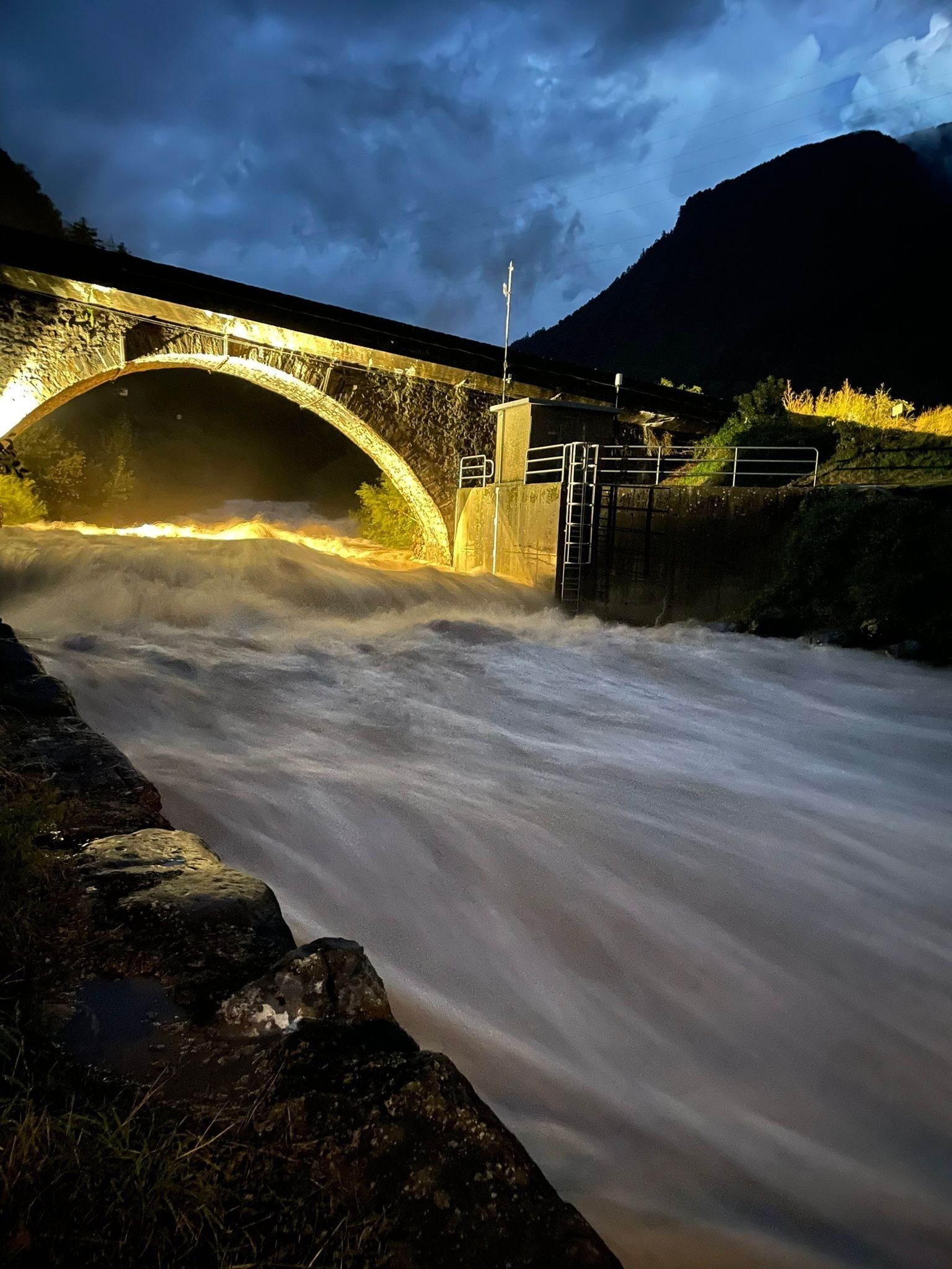 Un puente de piedra sobre un río caudaloso de noche, con una montaña al fondo.