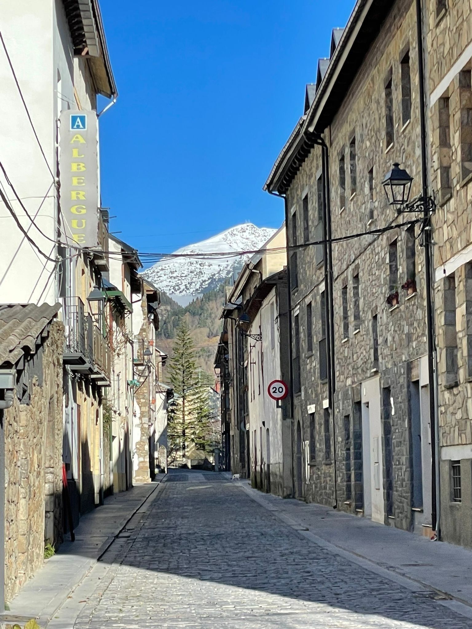 A narrow cobblestone street flanked by historic stone buildings with a mountain in the background.