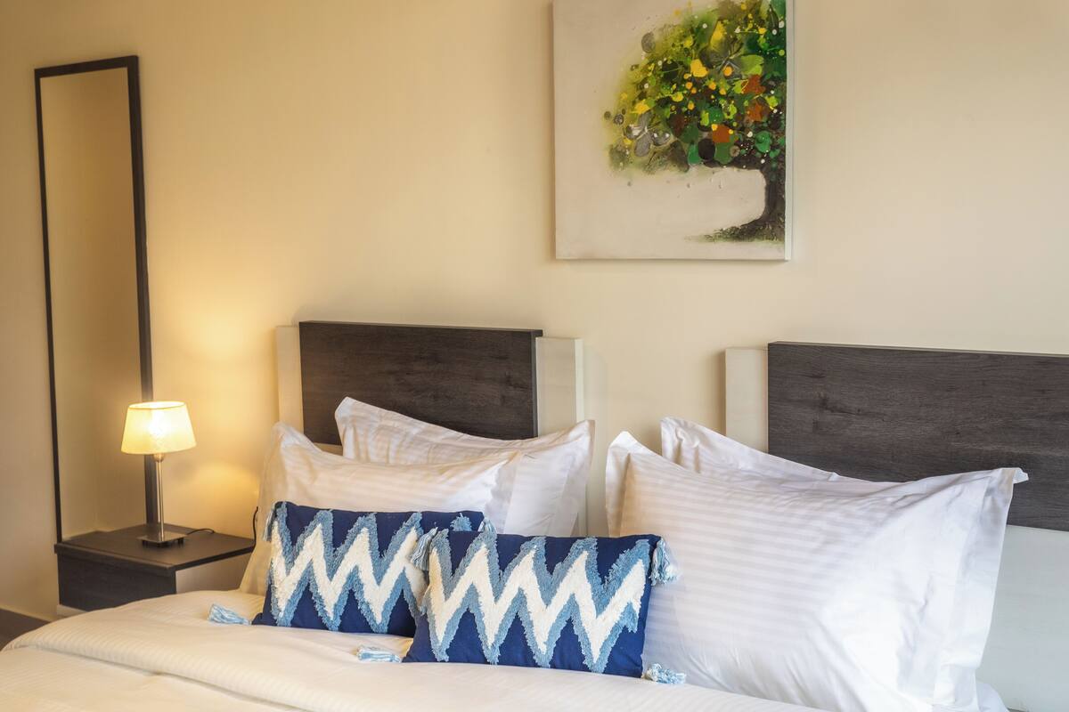 A neatly made bed with white and blue patterned pillows, a bedside lamp, and a painting of a tree on the wall.