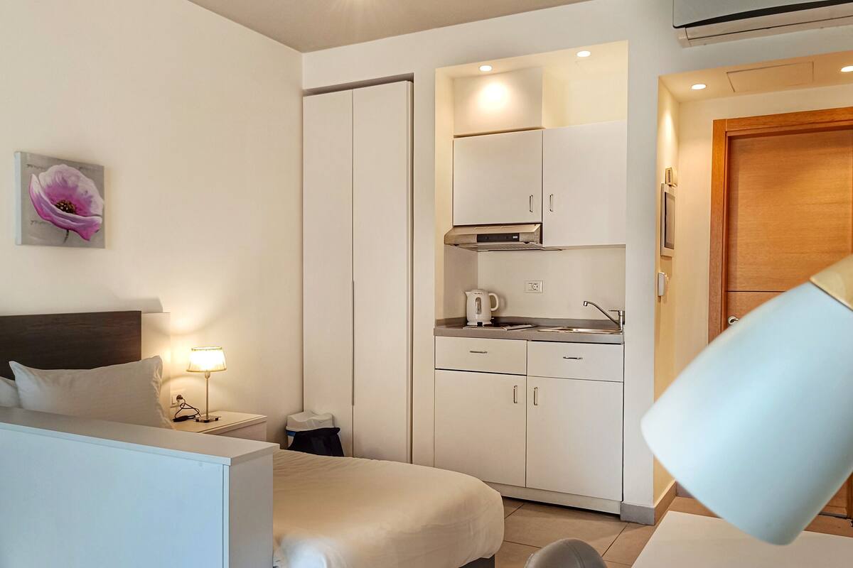 A hotel room with a bed, bedside table, lamp, and a kitchenette area with cabinets and a stove.